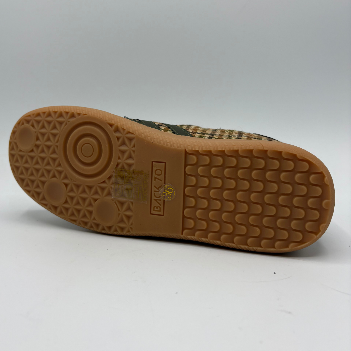 The outsole of a shoe with a brown textured rubber tread and a partially visible beige and black checkered fabric upper, shown on a light gray background.
