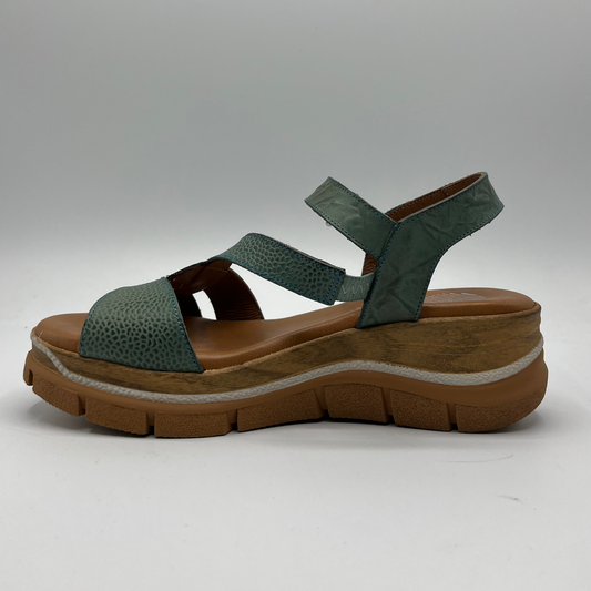 Left facing view of a leather sandal in shades of turquoise on the upper and an adjustable velcro strap and a thick wedge heel.