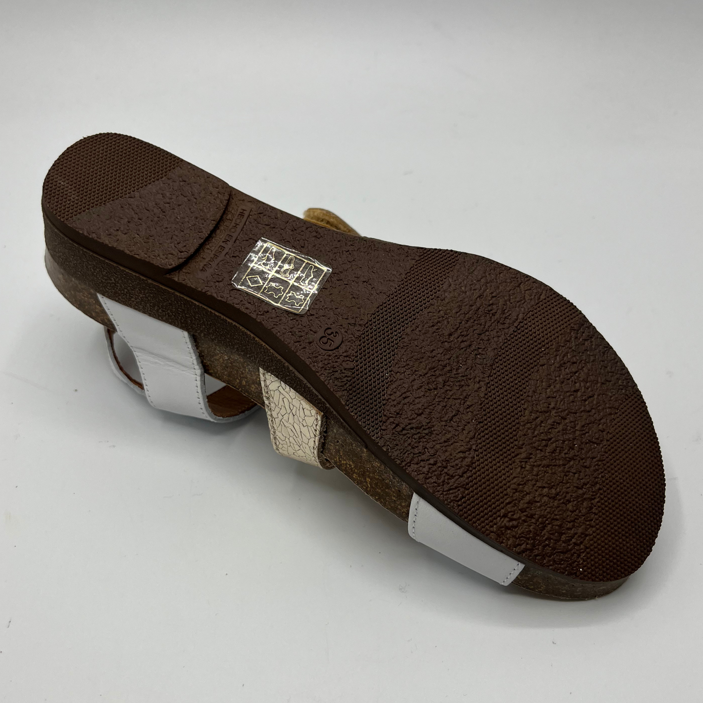 Bottom view of a leather sandal with white and champagne straps in a zigzag design. Two hook and loop adjustable straps and a low wedge heel.