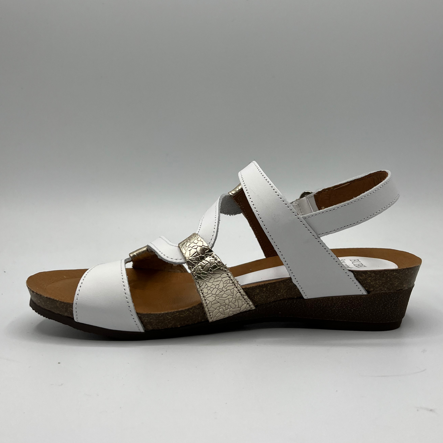 Left facing view of a leather sandal with white and champagne straps in a zigzag design. Two hook and loop adjustable straps and a low wedge heel.