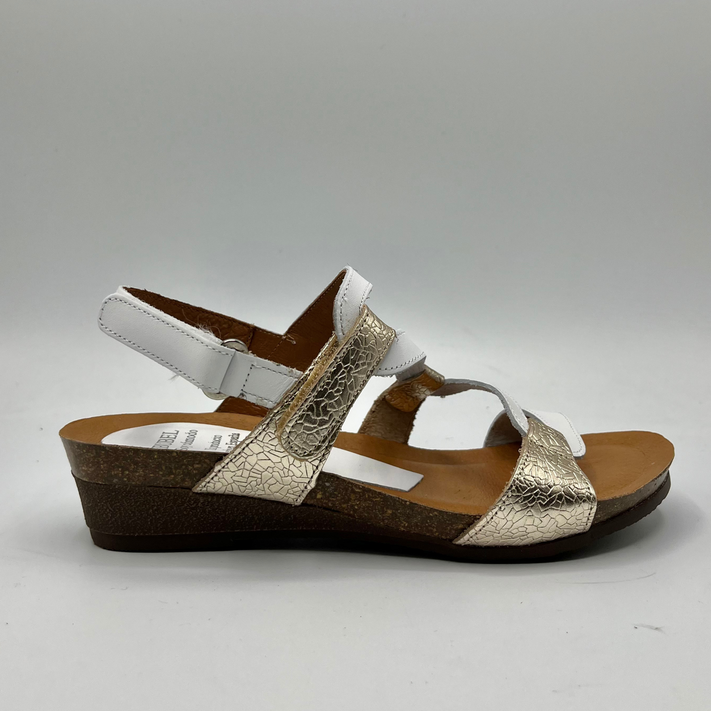 Right facing view of a leather sandal with white and champagne straps in a zigzag design. Two hook and loop adjustable straps and a low wedge heel.