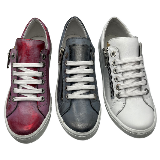 View of three sneakers in a row. The left one is metallic red, the middle is nickel and the right one is white. All crafted from leather with white laces and a side zipper closure