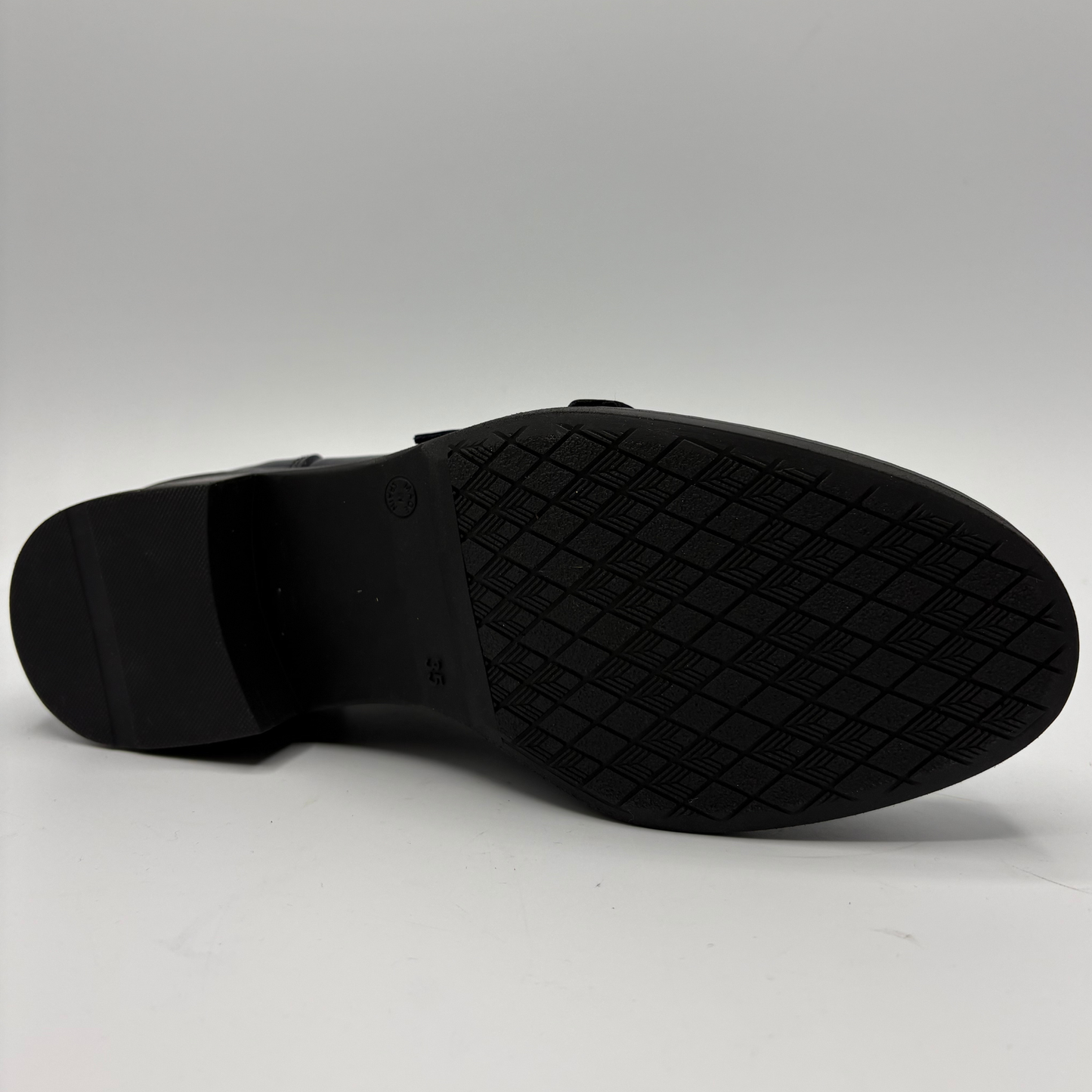 A close-up of a black shoe sole, showing a textured diamond-patterned rubber tread and a low block heel, photographed against a neutral light gray background.