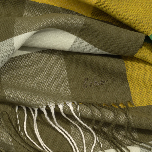 Close up view of a plaid scarf with yellow, taupe, cream and green colours and small tassels on each end.