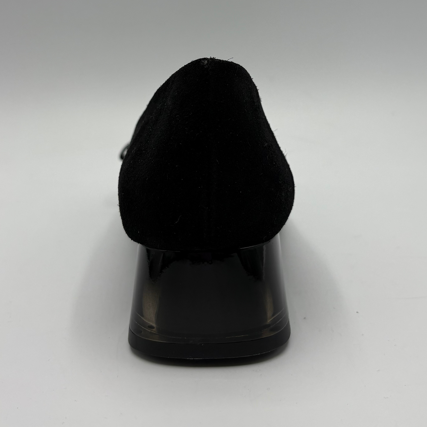 A black pointed-toe shoe shown from the back, featuring a textured black suede upper and a glossy black block heel, set against a plain light gray background.