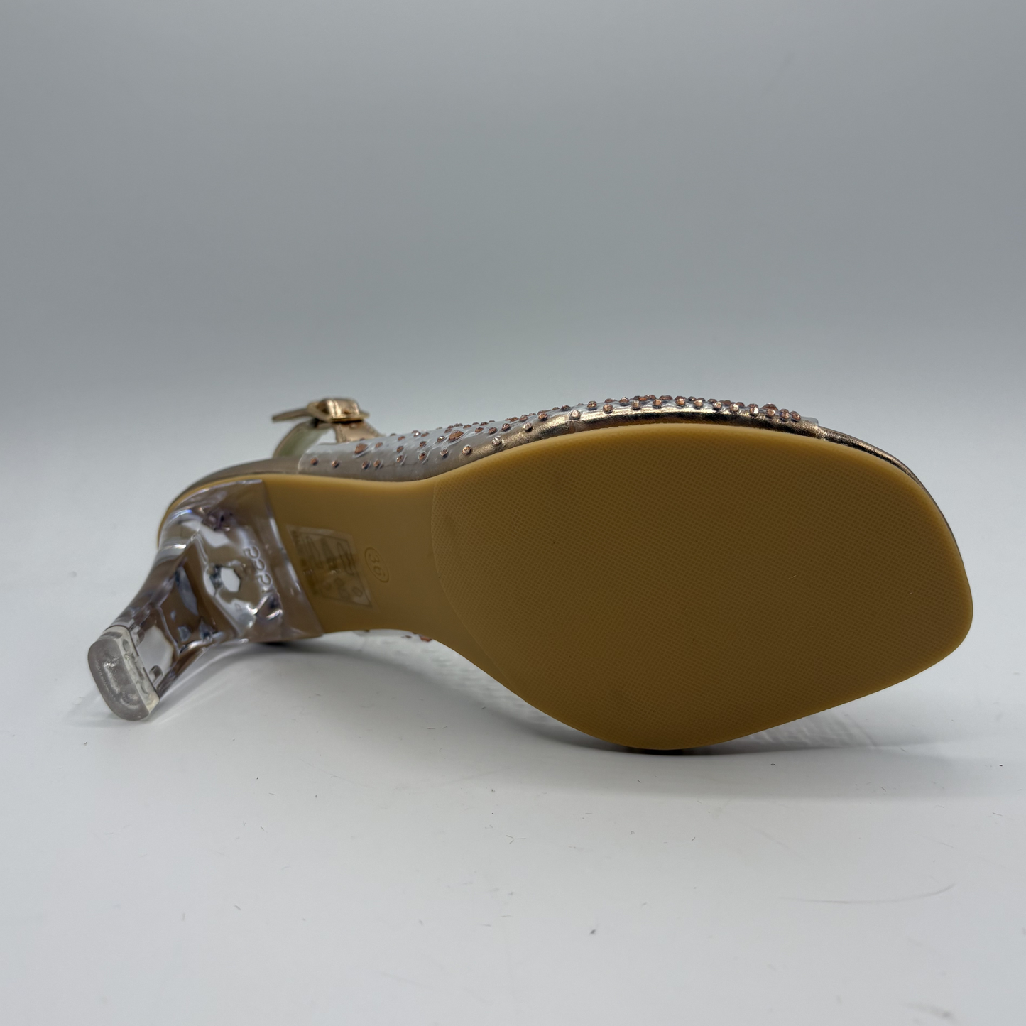 Back view of a sandal with clear strap and gold embellishments on a gray background