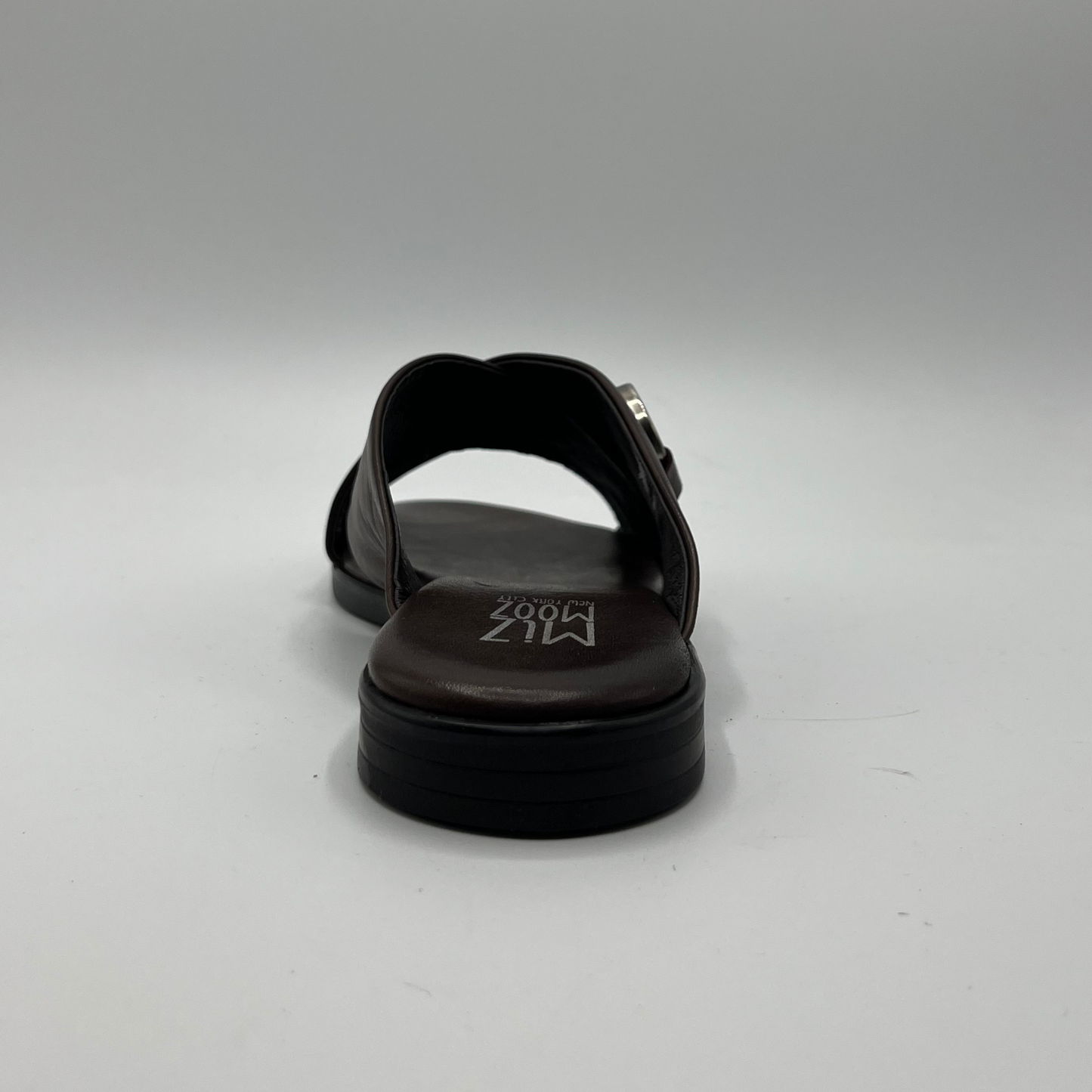 Back view of a brown leather slip on sandal with a cross strap design and low heeled outsole. Featuring a silver metal accent on the strap and a cushioned leather insole.