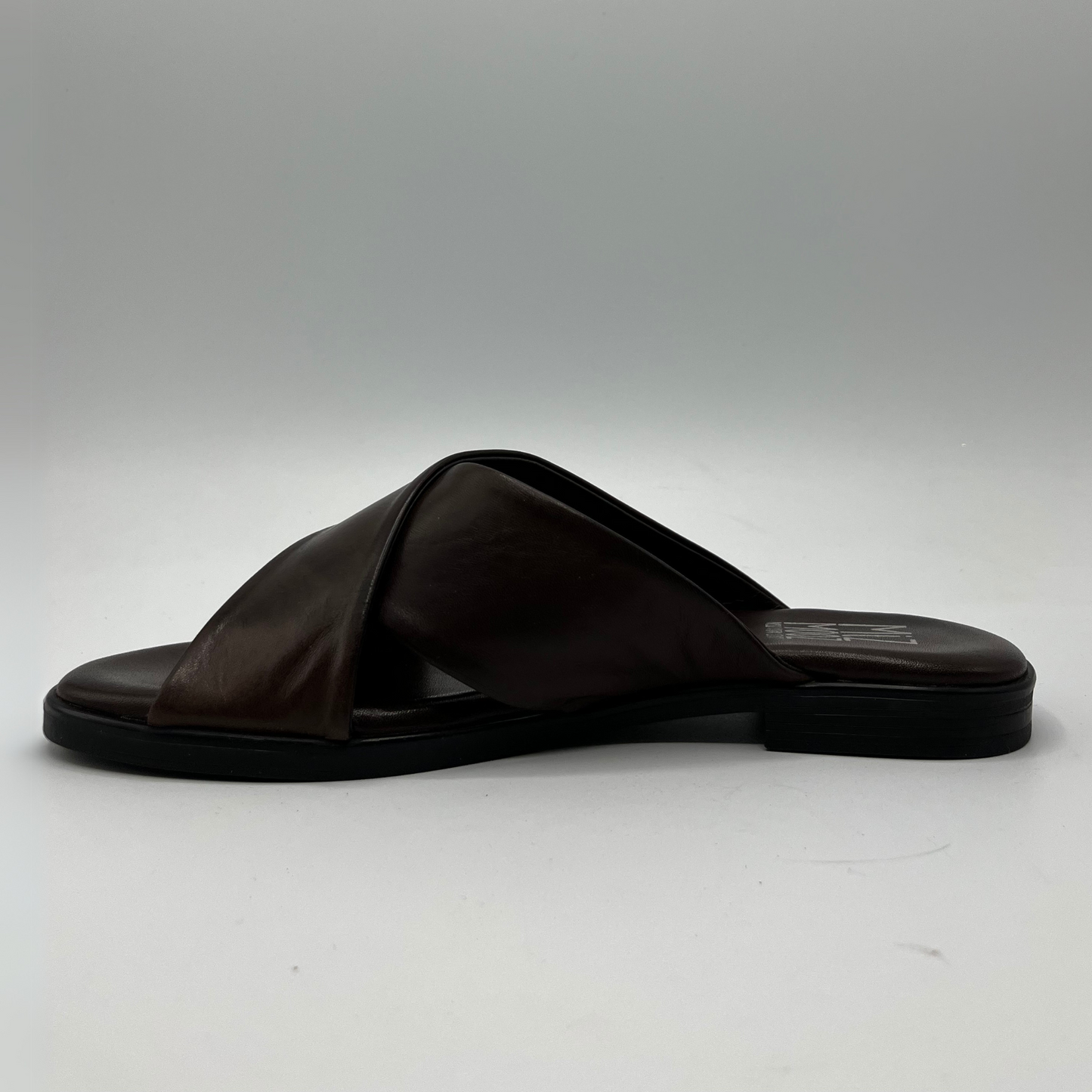 Left facing view of a brown leather slip on sandal with a cross strap design and low heeled outsole. Featuring a silver metal accent on the strap and a cushioned leather insole.