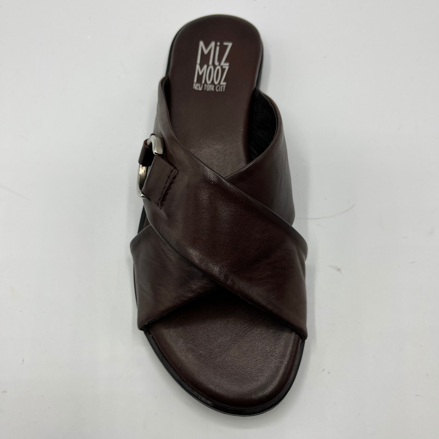 Top view of a brown leather slip on sandal with a cross strap design and low heeled outsole. Featuring a silver metal accent on the strap and a cushioned leather insole.