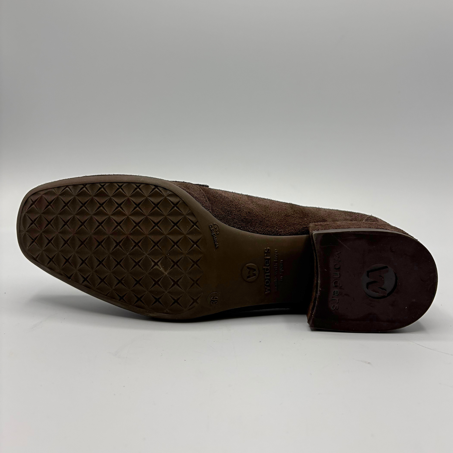 The outsole and heel of a brown suede shoe, showing a textured diamond-pattern tread and a dark brown rubber heel cap with a circular logo, photographed on a light gray background.