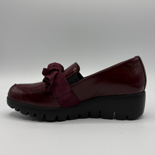 A single women's slip-on platform shoe in deep burgundy, featuring a glossy patent leather toe cap, a suede bow detail across the instep, and a chunky black lug sole, shown against a neutral gray background.