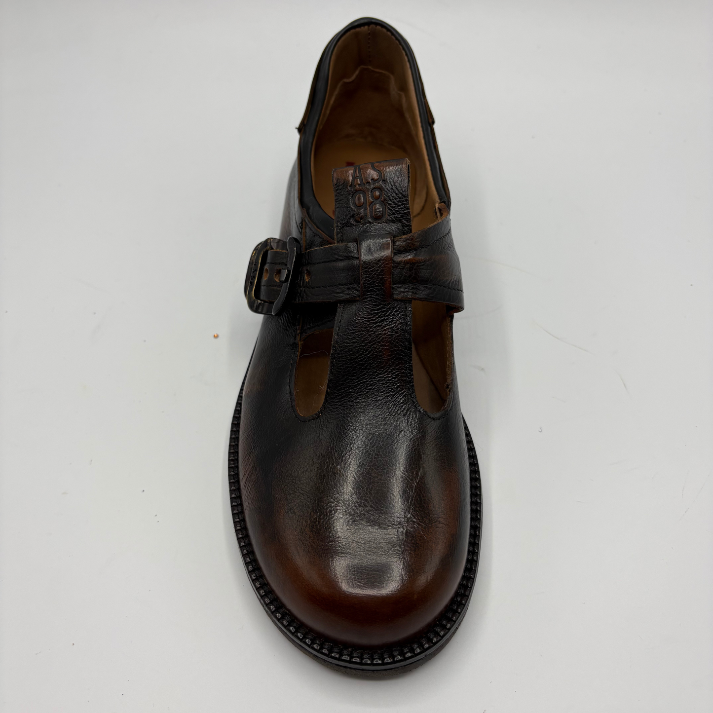 A dark brown leather T-strap Mary Jane shoe with a buckle strap, photographed from a top-down angle on a light gray background.