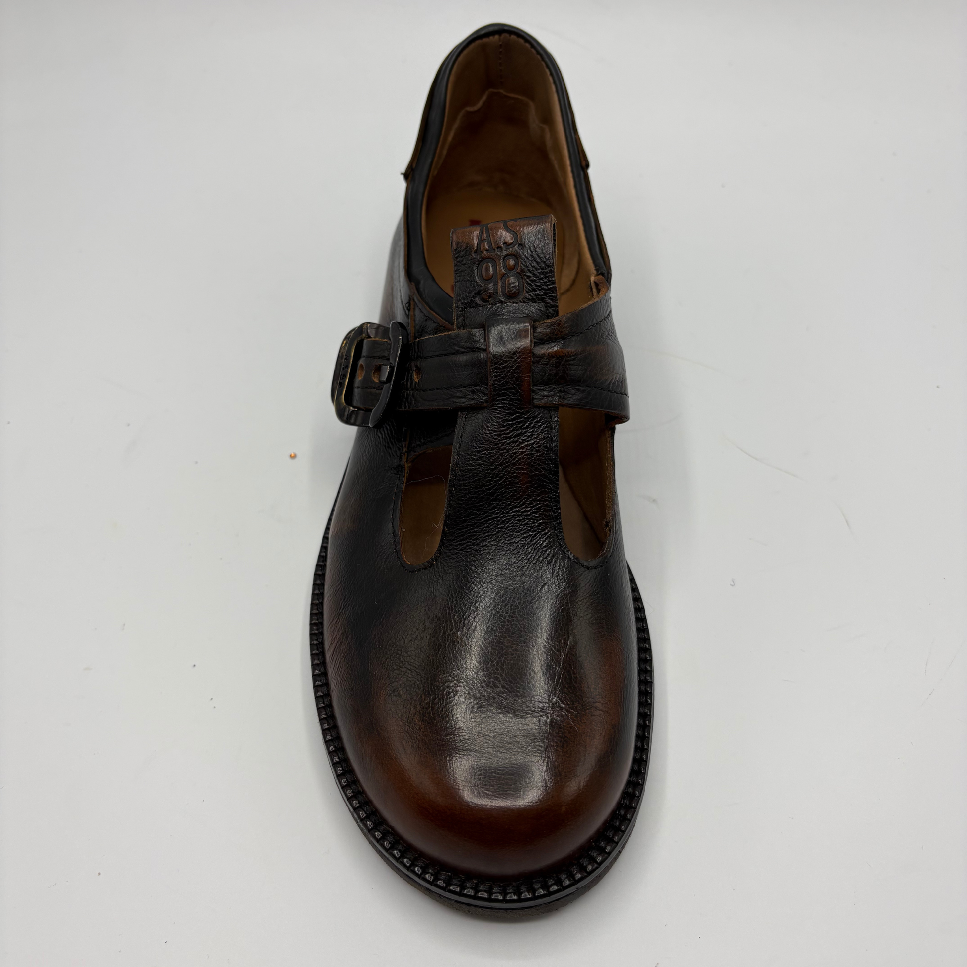 A dark brown leather T-strap Mary Jane shoe with a buckle strap, photographed from a top-down angle on a light gray background.
