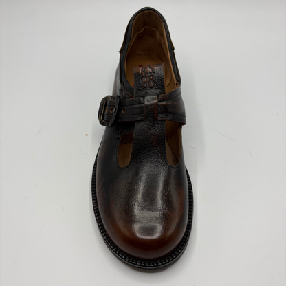 A dark brown leather T-strap Mary Jane shoe with a buckle strap, photographed from a top-down angle on a light gray background.