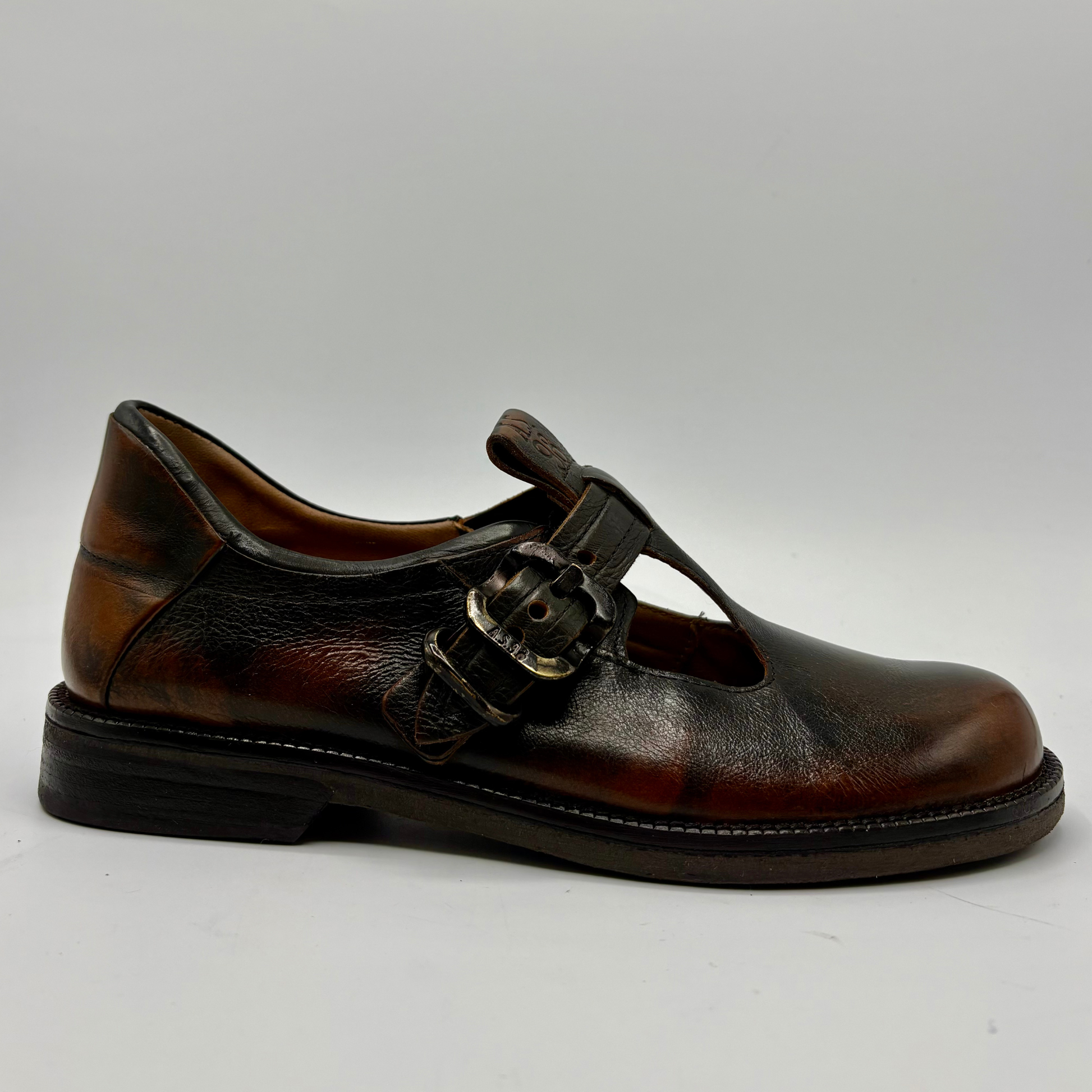 A single dark brown leather Mary Jane shoe with a T-strap and decorative buckle, shown from a side profile on a light gray background.