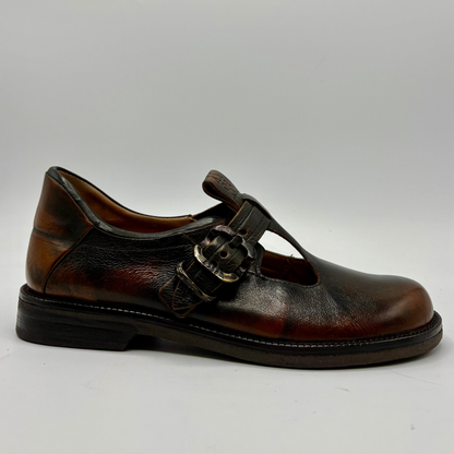 A single dark brown leather Mary Jane shoe with a T-strap and decorative buckle, shown from a side profile on a light gray background.