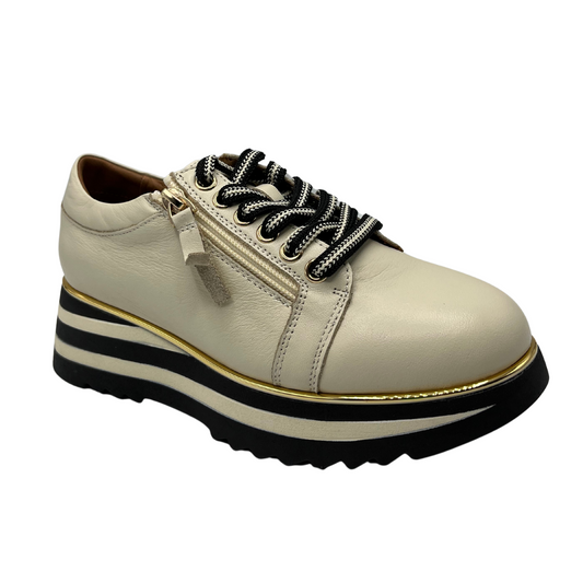 Angled view of cream coloured leather sneaker with a side zipper and lace up front. Thick striped platform sole with gold piping.