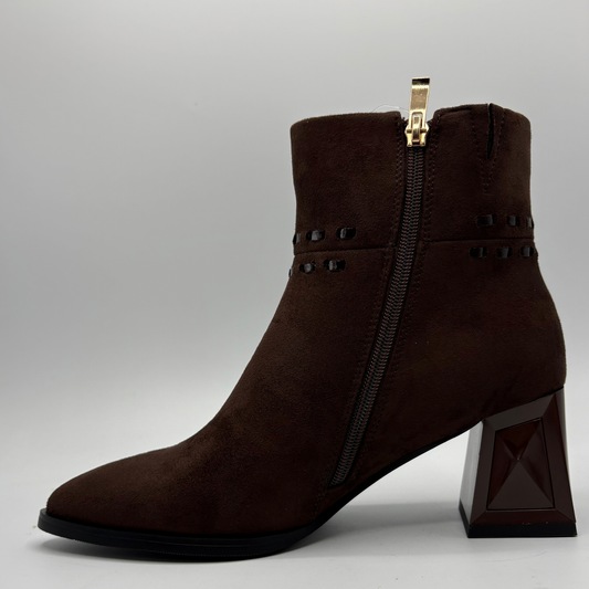 A dark brown suede ankle boot with a chunky geometric block heel, side zipper, and decorative studs, shown against a neutral gray background.