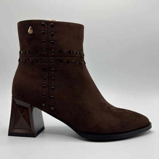 A dark brown suede ankle boot with a chunky geometric block heel, black studded detailing around the collar and side, and a small gold emblem on the side.