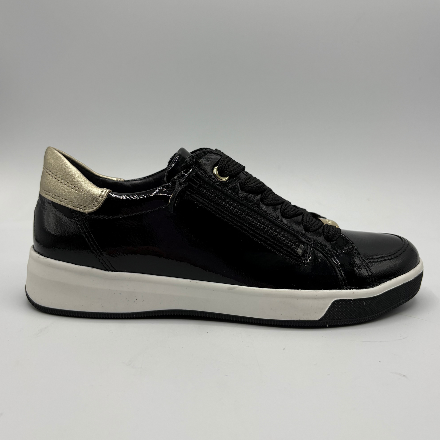 A black leather low-top sneaker with a gold metallic heel tab, black laces, a side zipper, and a thick white platform sole, shown from a side profile on a neutral gray background.