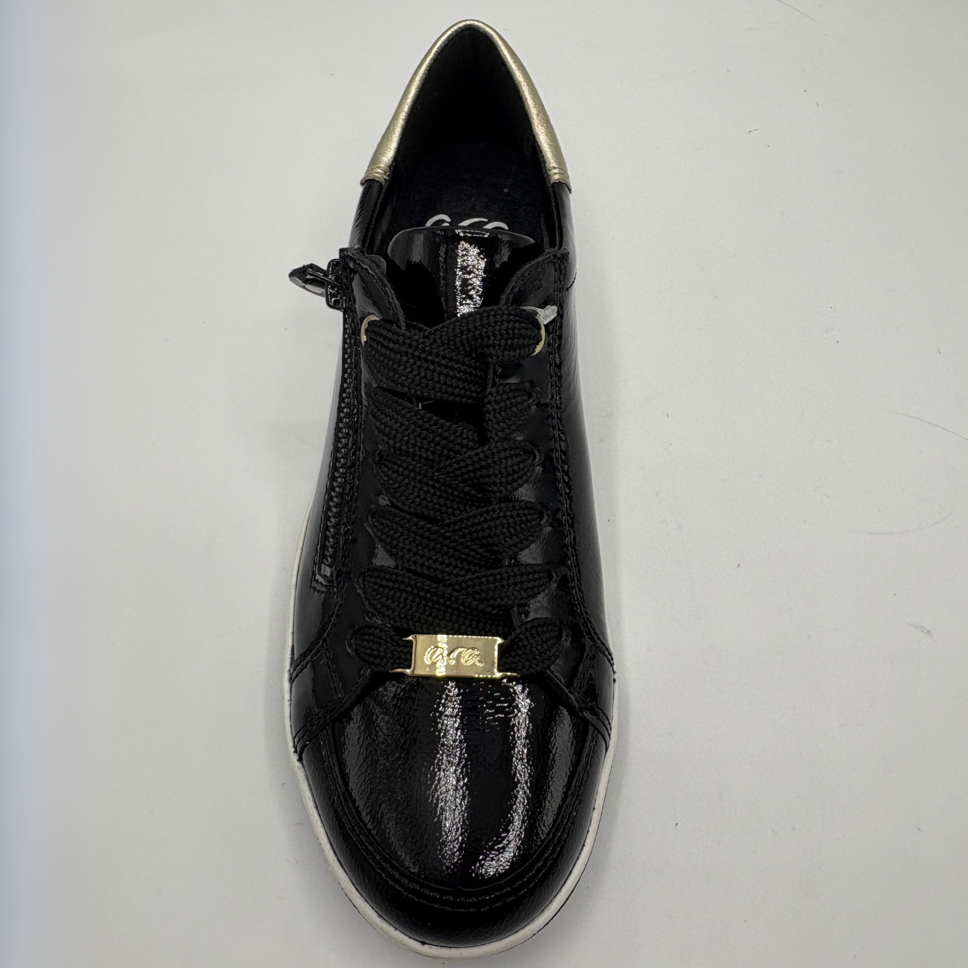 A black low-top sneaker with a glossy patent leather toe cap, black laces, and a gold metal accent plate on the front, photographed from a top-down angle on a light gray background.