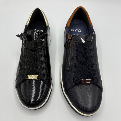 A pair of low-top sneakers shown from the front and side on a light gray background. The shoes feature black laces, a side zipper on the right shoe, a metallic gold logo plate on the front, and a white midsole.