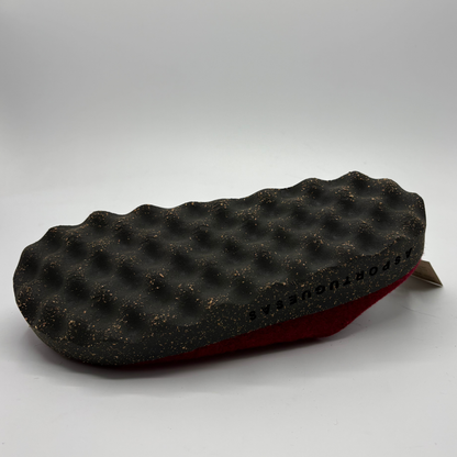 A close-up of a shoe sole showing the textured black outsole with small gold flecks with a red upper visible underneath, photographed on a light gray studio background.