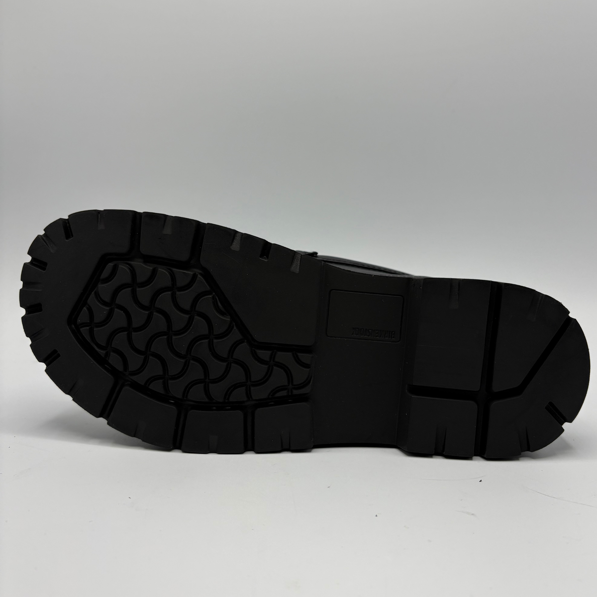A close-up view of a black rubber shoe sole, showing the tread pattern and lug design on a light gray background.