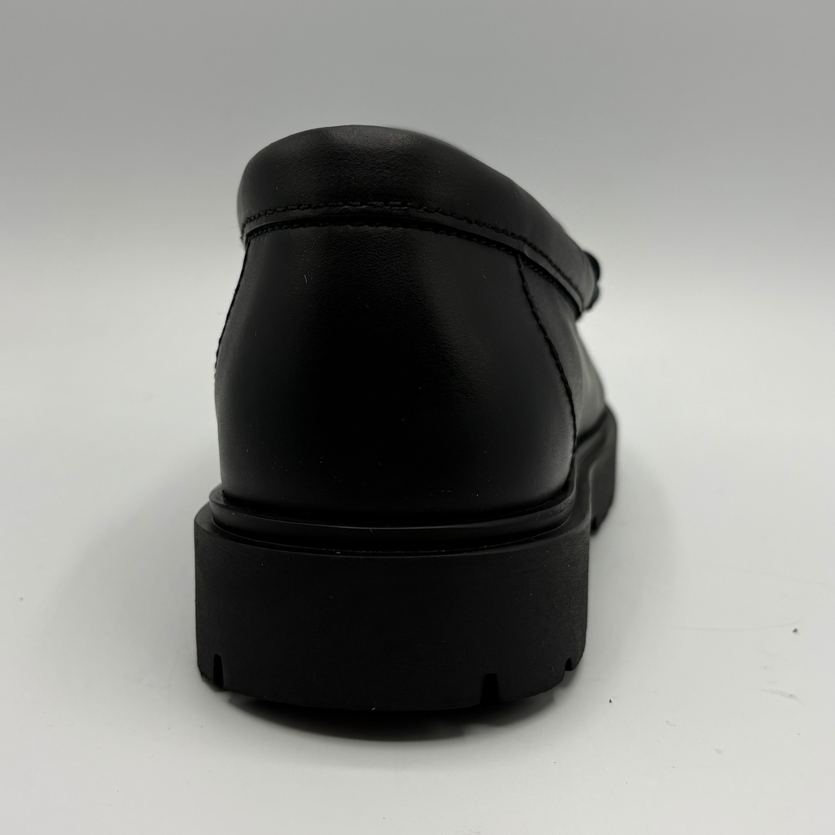 A black leather loafer shown from a rear three-quarter angle, featuring a thick black platform sole, photographed on a neutral light gray background.