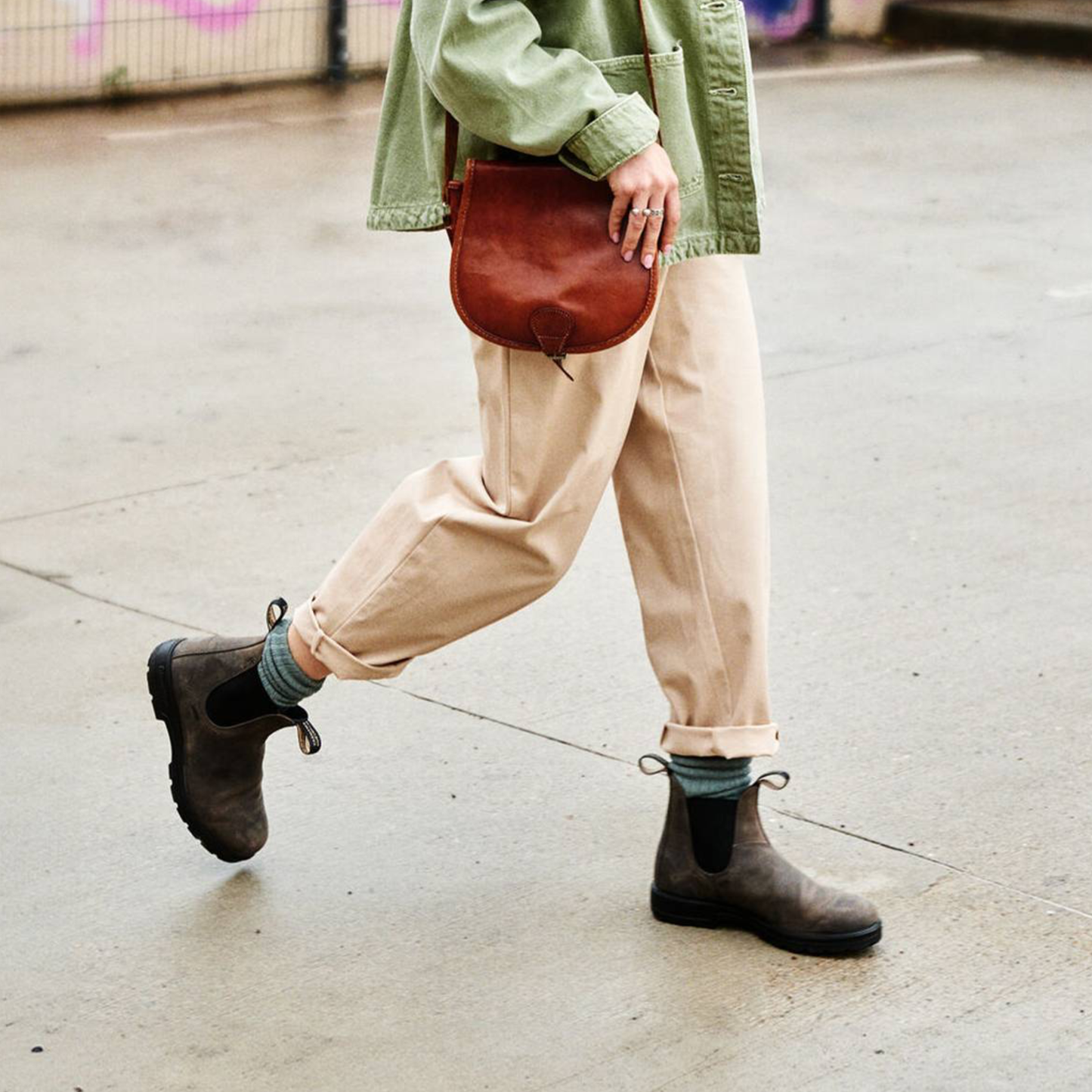 Person wearing beige pants, green jacket, and brown boots with a brown leather bag on a street.