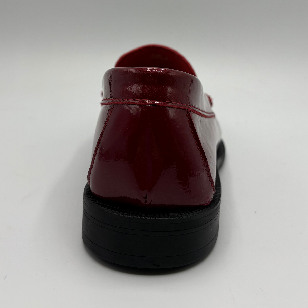 A glossy patent leather loafer shown from a rear three-quarter angle, featuring a deep burgundy red upper with matching red stitching and a chunky black sole, set against a plain light gray background.