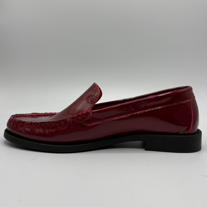 A single glossy burgundy leather loafer with black stitching and a black sole, shown in a side profile against a neutral gray background.