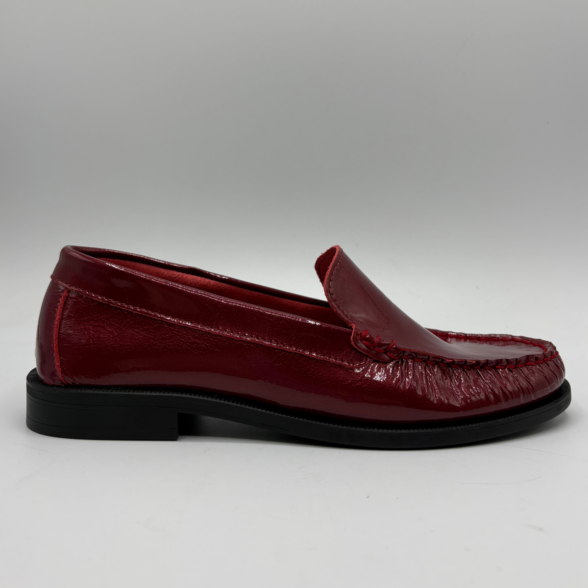A single glossy burgundy leather moccasin loafer with a black sole, shown in a side profile against a neutral gray background.