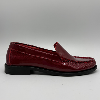 A single glossy burgundy leather moccasin loafer with a black sole, shown in a side profile against a neutral gray background.