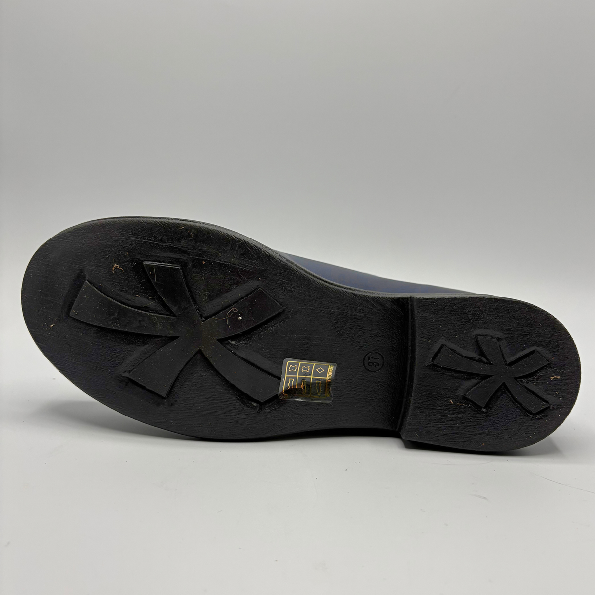 The black rubber outsole of a shoe, showing a distinctive four-pointed star-like tread pattern on the front and heel, with a small gold brand tag in the center.