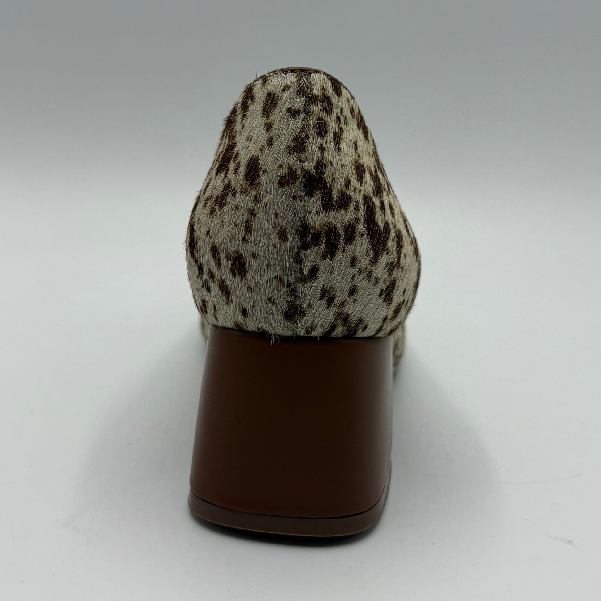 A single women's pump shoe shown from the back, featuring a brown and beige animal-print hair-on-hide upper and a solid dark brown block heel, set against a neutral gray studio background.
