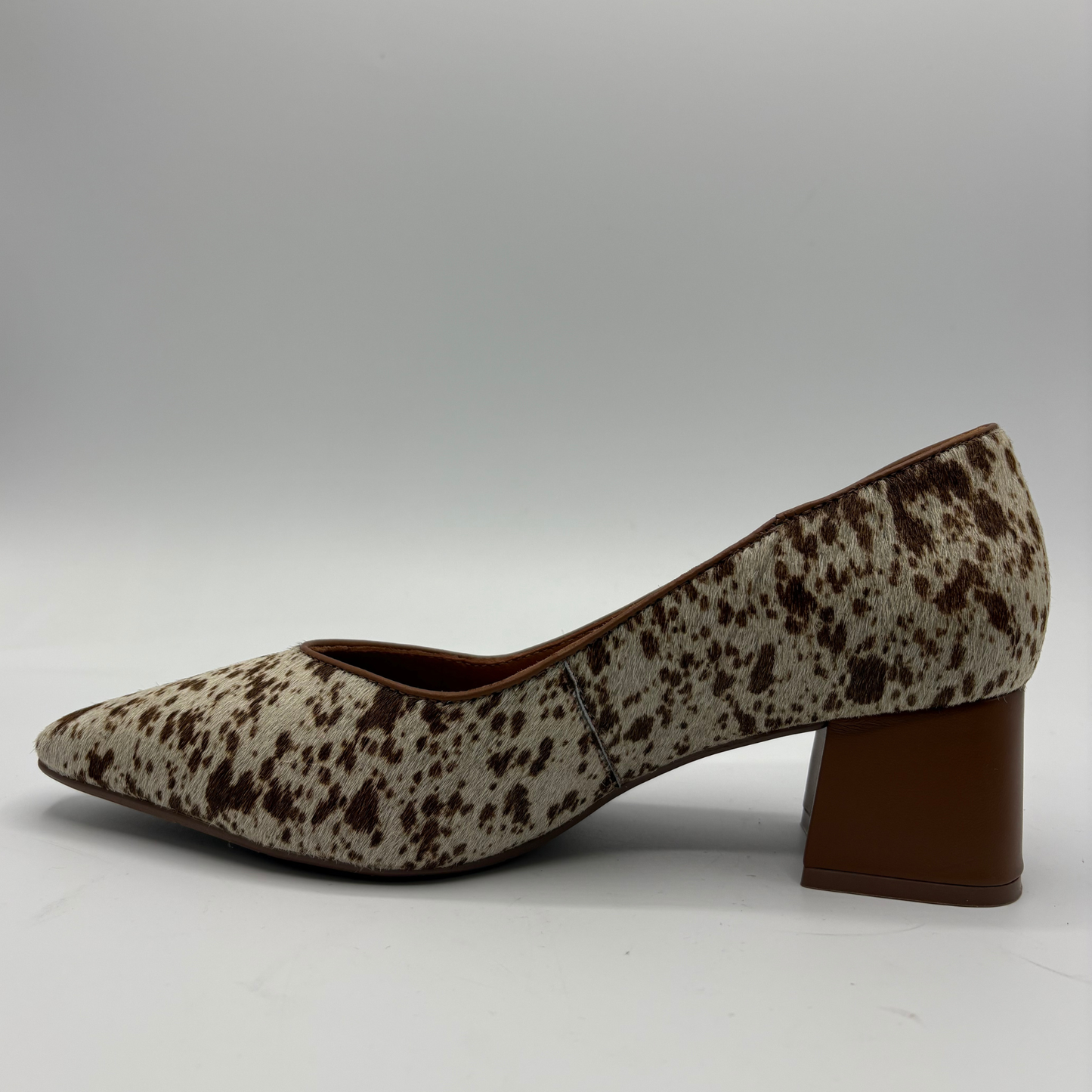 A single women's pump shoe with a brown and beige cow-print pattern, featuring a pointed toe and a brown block heel, shown from a side profile on a light gray background.