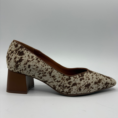 A single women's pump shoe with a brown and beige cow-print pattern, featuring a pointed toe and a chunky brown block heel, shown against a neutral gray background.