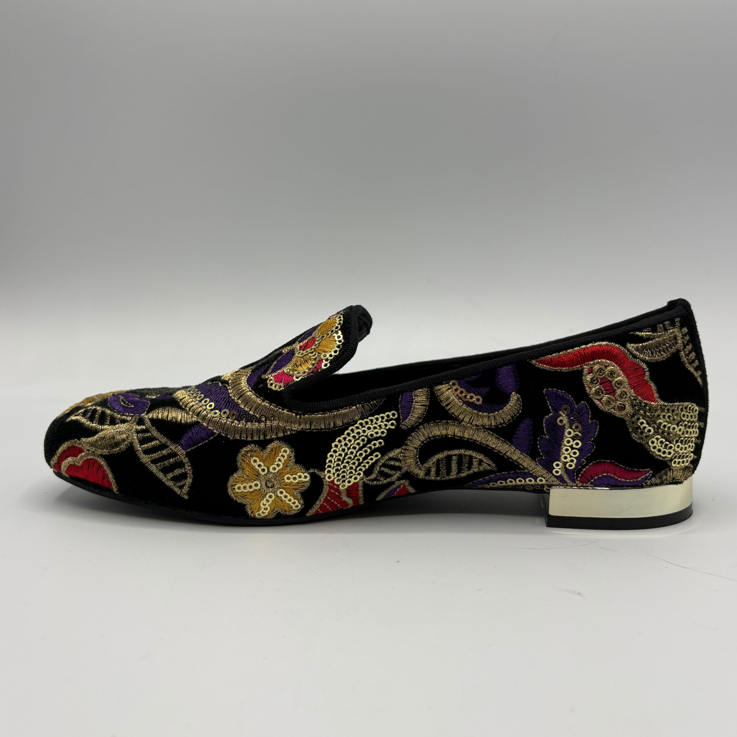 A single black velvet flat shoe decorated with intricate multicolor embroidery in gold, purple, red, and beige, shown from a side profile on a neutral gray background.