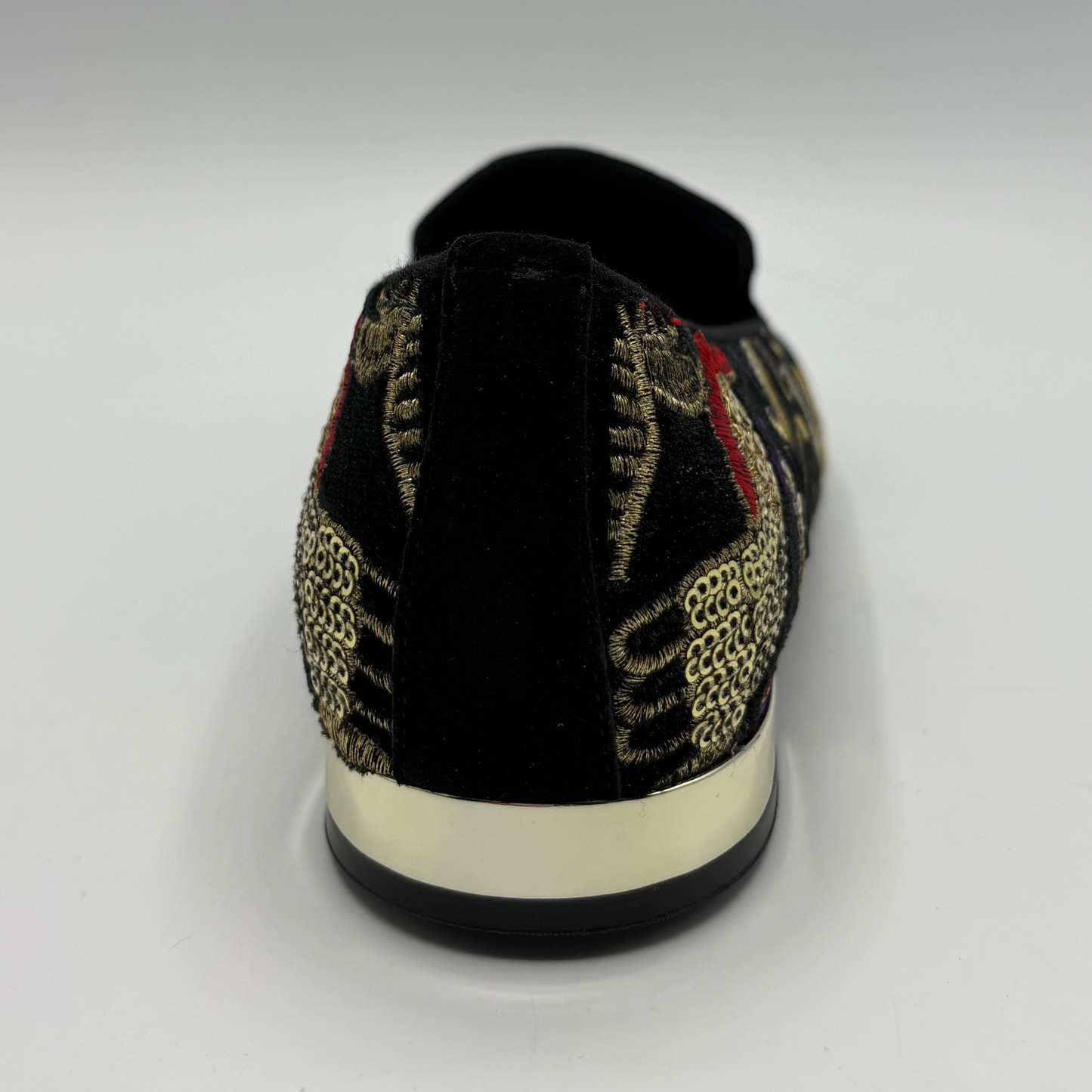 A single slip-on shoe shown from the back on a light gray background. The shoe features a black velvet heel and upper, a black and gold woven patterned side, a red accent stripe, and a thick cream-colored midsole.