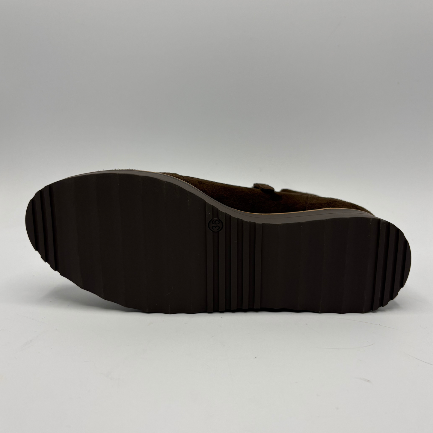 The outsole of a dark brown shoe, showing a ribbed tread pattern and a small circular logo embossed in the center, photographed against a neutral gray studio background.