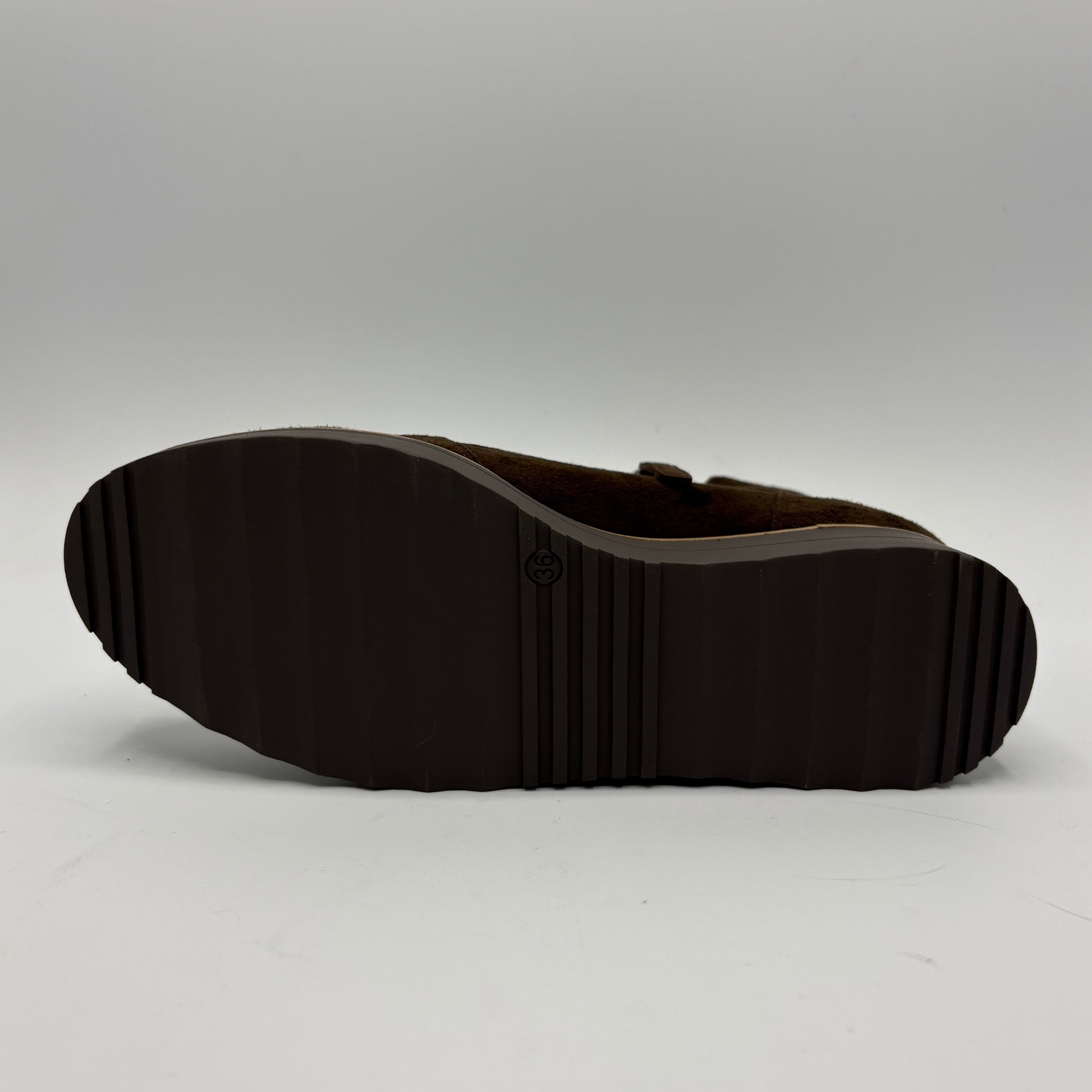 The outsole of a dark brown shoe, showing a ribbed tread pattern and a small circular logo embossed in the center, photographed against a neutral gray studio background.