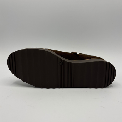 The outsole of a dark brown shoe, showing a ribbed tread pattern and a small circular logo embossed in the center, photographed against a neutral gray studio background.