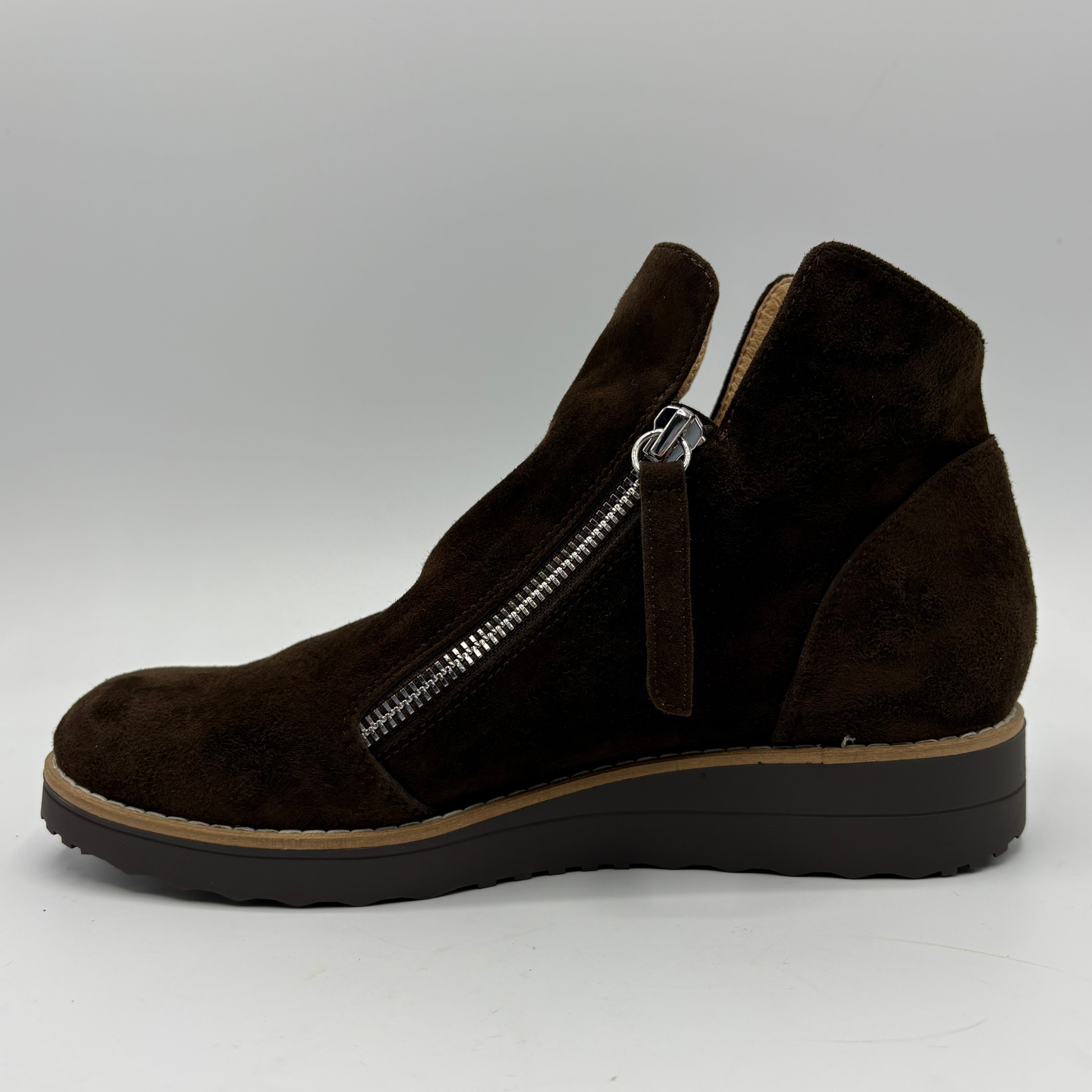 A dark brown suede ankle boot with a side zipper, a pull tab, and a wedge heel, shown in profile against a light gray background.