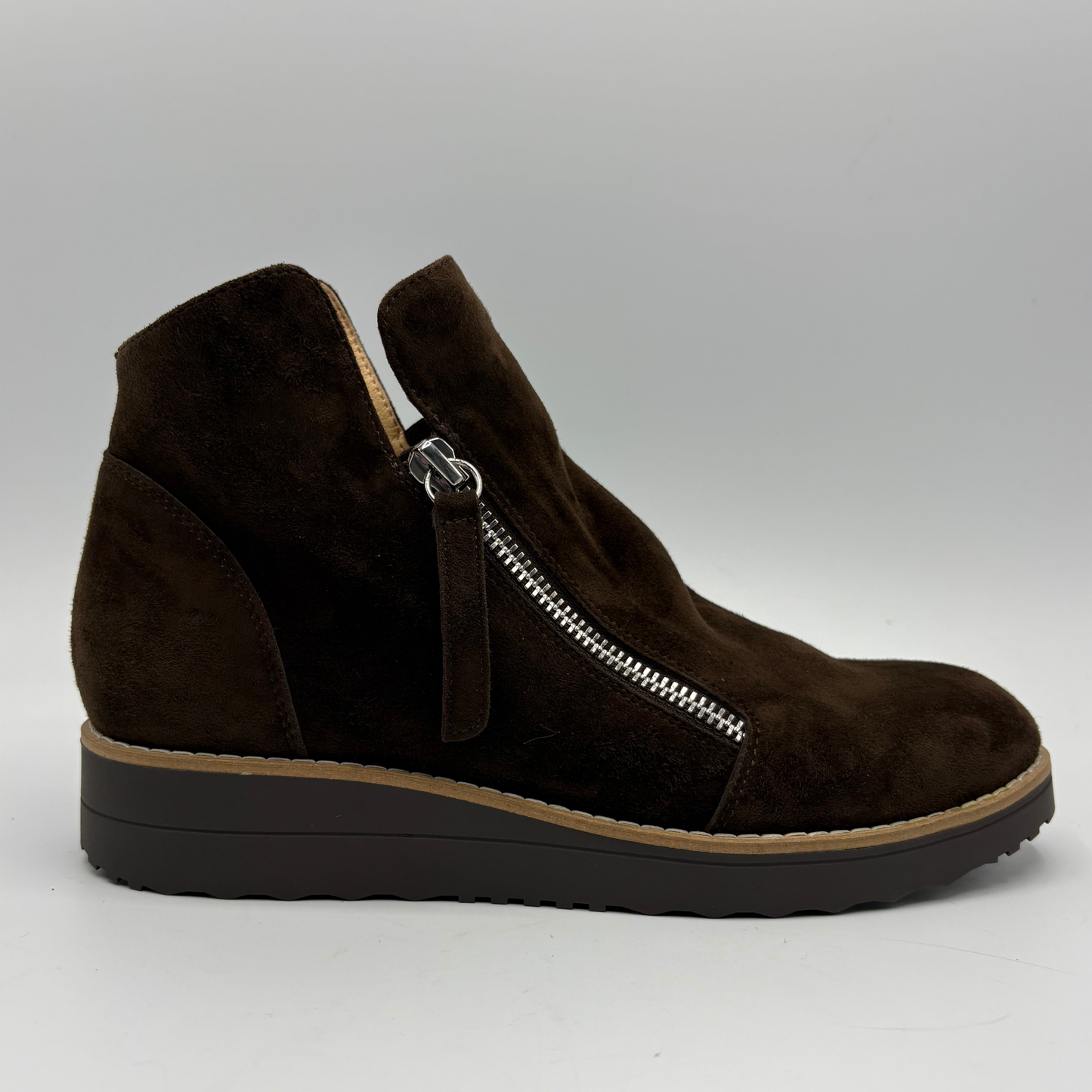 A dark brown suede ankle boot with a side zipper, a low wedge heel, and a tan outsole, shown from a side profile view against a neutral gray background.