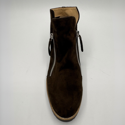 A dark brown suede ankle boot with silver zipper details on the sides, photographed from a top-down perspective on a light gray background.