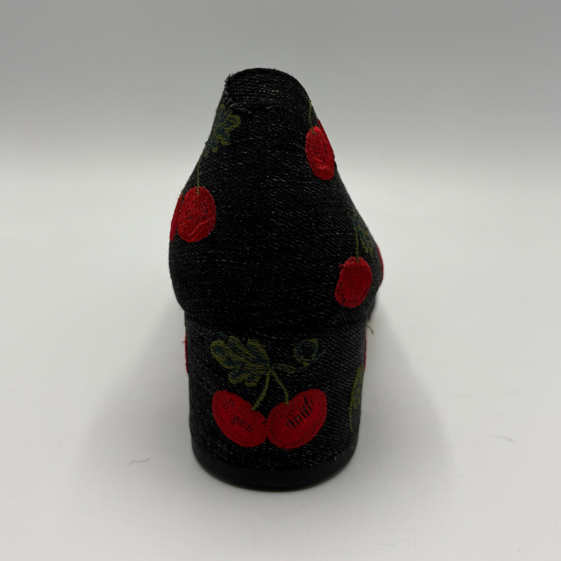 A black fabric-covered shoe heel or sole section with embroidered red cherries and green leaves, photographed against a plain light gray studio background.