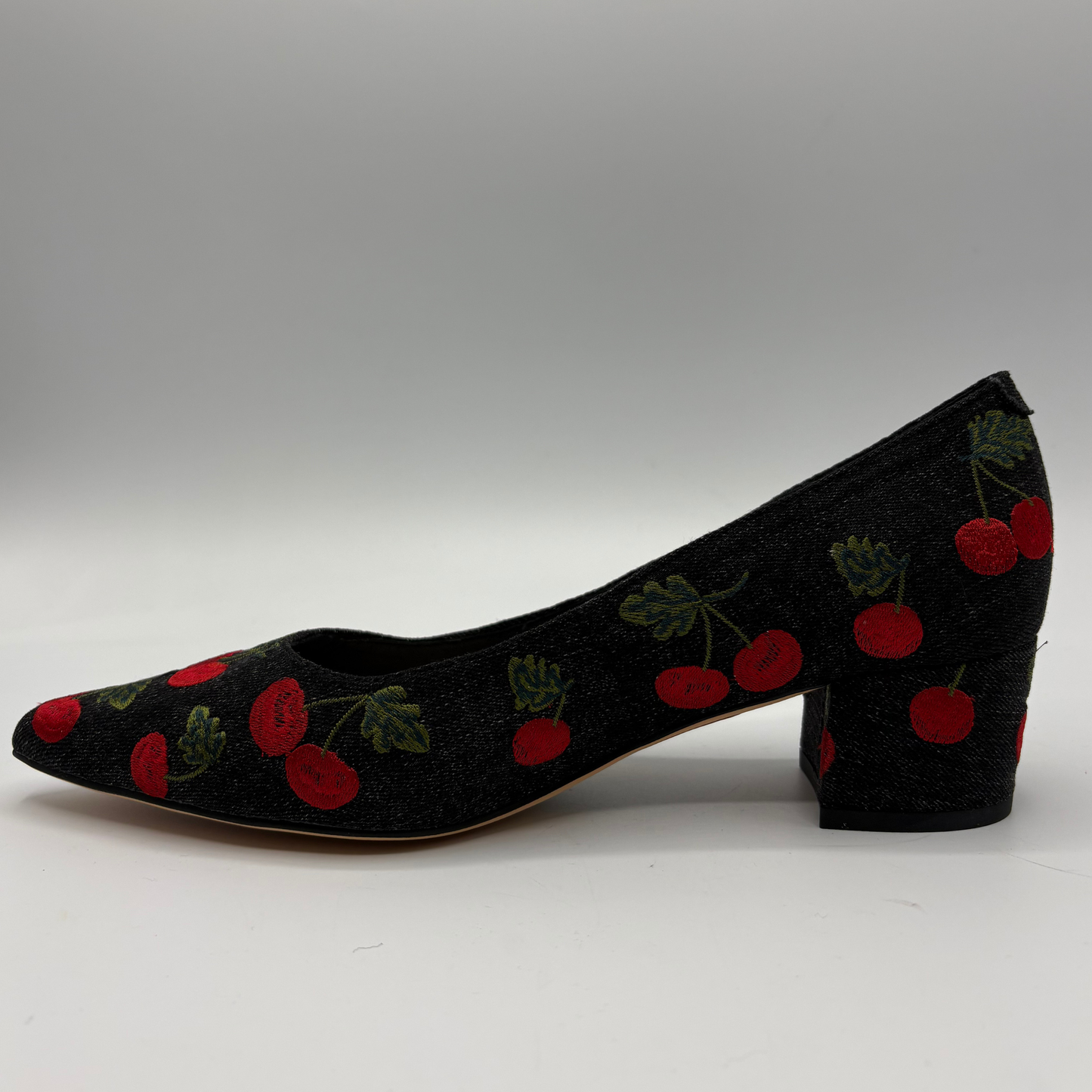 A single black pointed-toe pump with a block heel, covered in a red cherry and green leaf pattern, shown against a neutral gray background.