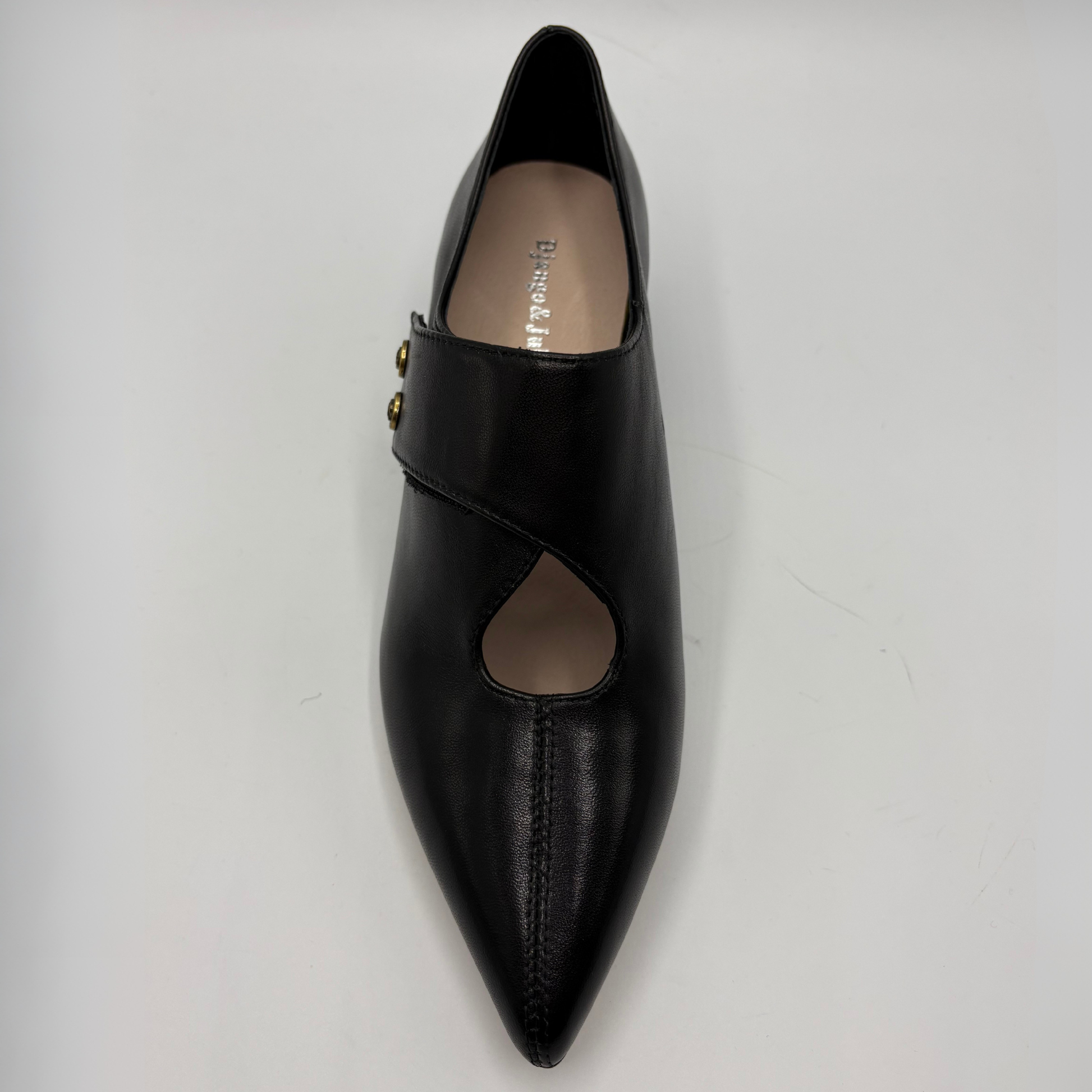 A single black leather pointed-toe flat shoe with a wrap-around strap and two small gold-tone decorative studs, photographed from a top-down angle on a light gray background.