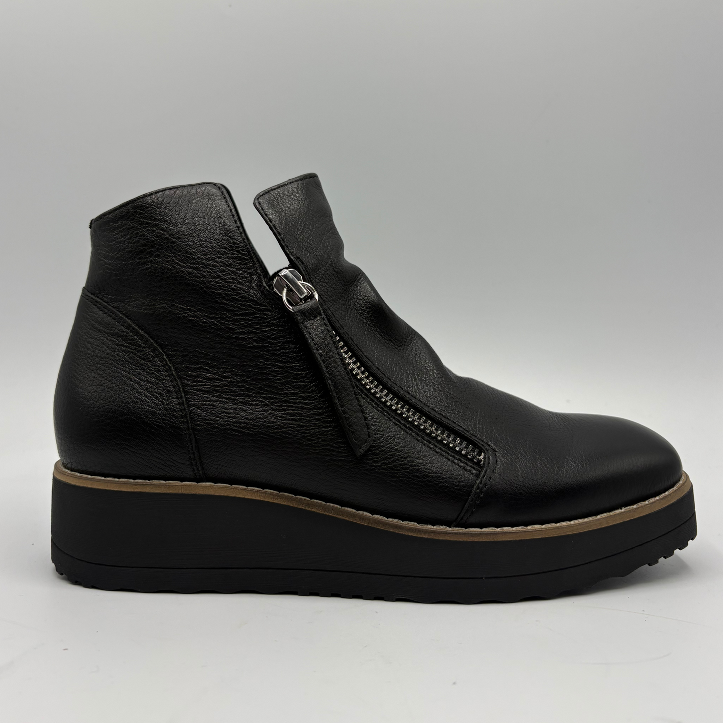 A black leather ankle boot with a side zipper, a chunky black platform sole with a tan mid-layer, and a silver-tone metal buckle, shown against a neutral gray background.